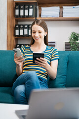 Fototapeta premium Woman sitting on sofa using mobile phone to calculate household expenses, hand holding account book for income-expense.