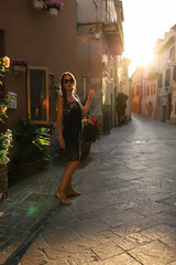 Una giovane ragazza bruna dai capelli fluenti in un tubino nero per le strade dei vicoli d'Italia al tramonto con vasi di fiori alle pareti