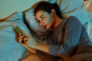 Top view portrait of smiling adult woman using smartphone in bed at night reading text messages and...