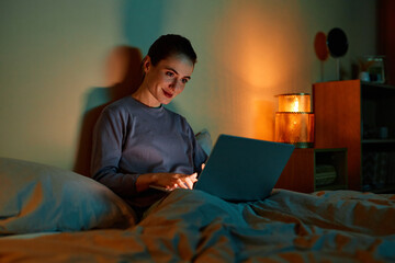 Portrait of smiling adult woman using laptop in bed at night, copy space