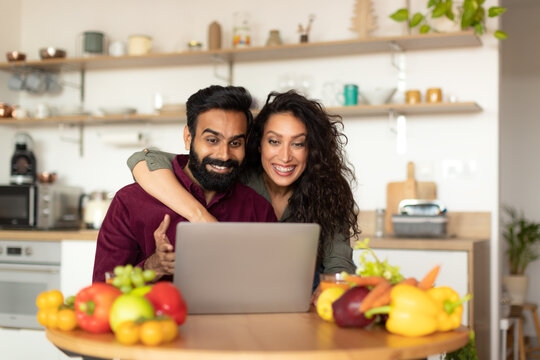 Happy arab spouses using laptop at kitchen, cheerful husband and wife shopping online or ordering food delivery