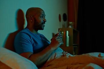 Side view portrait of smiling black man watching TV in bed at night staying up late, copy space