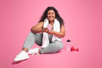 Positive black chubby woman relaxing with smartphone after workout, checking sports videos online