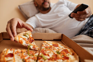 Closeup of adult man taking slice of pizza while enjoying lazy weekend at home and watching TV, copy space