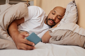 Portrait of adult black man lying in bed at home and using smartphone scrolling social media