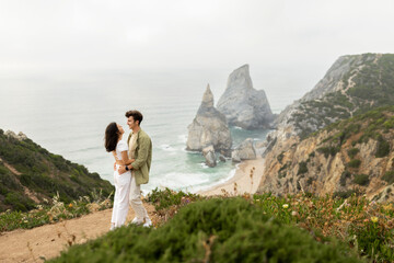 Couple in love having romantic date, embracing, standing on rocky cliff by the ocean with breathtaking view at coastline
