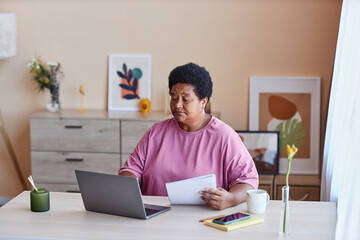 Serious businesswoman with document sitting by desk in front of laptop in home office and looking through online information on screen