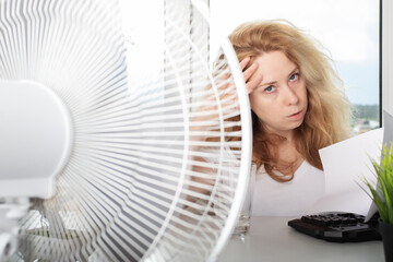 Woman blowing a fan to prevent overheating and sweating in the office, Adapting to the heat