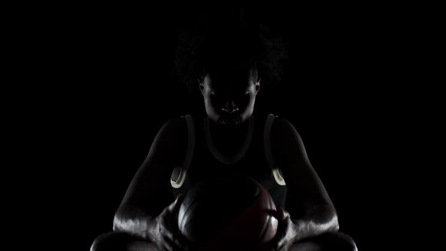 Basketball player silhouette sitting on a bench holding a ball. Side lit concentrated african american sports man against black background.