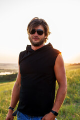 a young blond guy in a black T-shirt and sun goggles is standing on the grass enjoying the rays of the setting sun