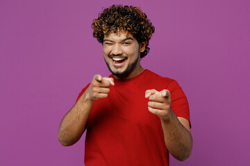 Young cheerful smiling happy Indian man he wearing red t-shirt casual clothes pointing index finger camera on you wink blik eye isolated on plain purple background studio portrait. Lifestyle concept.