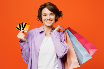 Young happy woman wearing purple shirt casual clothes hold in hand discount cards paper package bags after shopping isolated on plain orange color background studio. Black Friday sale buy day concept.