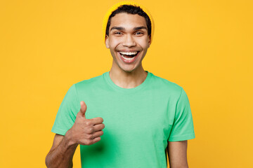 Young satisfied cheerful man of African American ethnicity he wears casual clothes green t-shirt hat show thumb up like gesture isolated on plain yellow background studio portrait. Lifestyle concept.