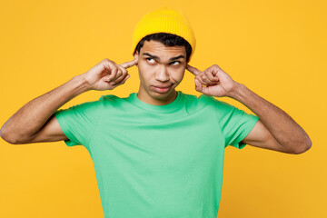 Naklejka premium Young sad man of African American ethnicity he wearing casual clothes green t-shirt hat cover ears with hands fingers do not want to listen scream isolated on plain yellow background studio portrait.