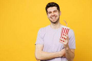 Young smiling cheerful fun happy caucasian man wear light purple t-shirt casual clothes hold in hands soda pop cola fizzy water isolated on plain yellow background studio portrait. Lifestyle concept.