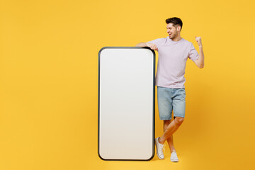 Full body young excited smiling man wearing light purple t-shirt casual clothes big huge blank screen mobile cell phone smartphone with area doing winner gesture isolated on plain yellow background.