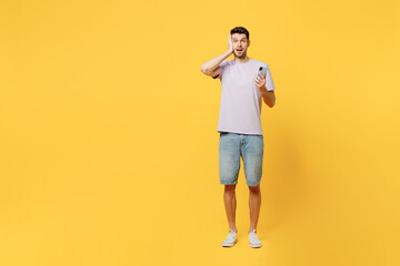Fototapeta na wymiar Full body young sad scared frightened amazed man he wearing light purple t-shirt casual clothes hold head use mobile cell phone isolated on plain yellow background studio portrait. Lifestyle concept.