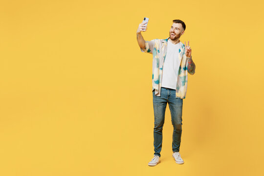 Full body young man he wear blue shirt white t-shirt casual clothes doing selfie shot on mobile cell phone post photo on social network show v-sign isolated on plain yellow background studio portrait.