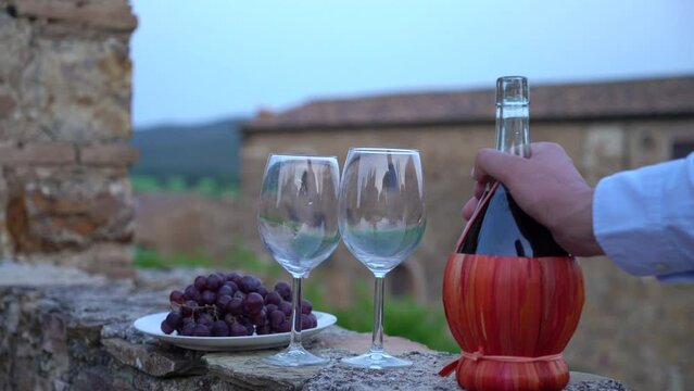 Man pouring red wine into a glass from the typical chianti wine bottle fiasco against the backdrop of the Tuscany landscape