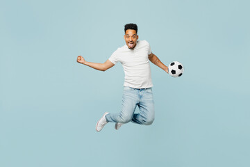Fototapeta premium Full body overjoyed young man fan wears t-shirt do winner gesture cheer up support football sport team hold soccer ball jump high watch tv live stream isolated on plain pastel blue color background.