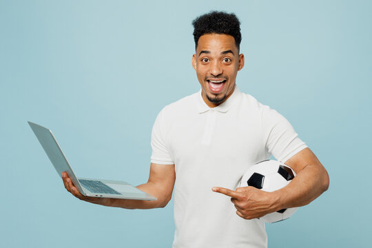Young IT man fan wears basic t-shirt cheer up support football sport team hold in hand use work on laptop pc computer soccer ball watch tv live stream isolated on plain pastel blue color background.