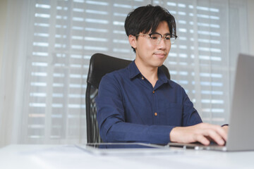Happy young adult Asian businessman, male businessperson using computer laptop on desk at office. Professional entrepreneur business man wear eyeglasses, sitting to working, planning on workplace. - 619086731