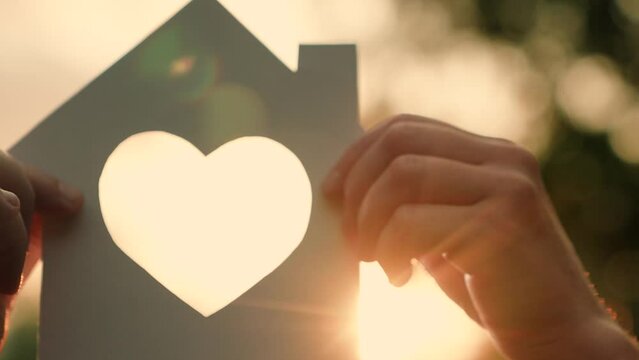 Hands holding paper house, sun shines in window. Family hands, paper house closeup at sunset. Symbol home, family comfort, happiness. Symbol heart. Concept of dream to buy house. Real estate insurance