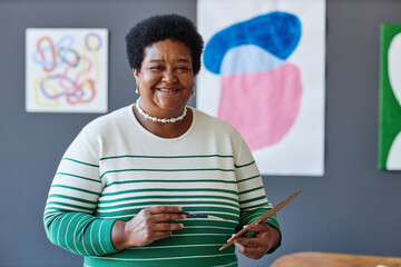 Mature smiling teacher of arts with watercolor palette looking at camera while standing in studio...