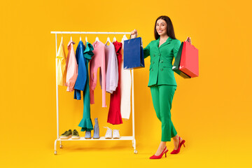 Great sales. Excited shopaholic lady holding colorful shopping bags, happy about new clothes standing near clothing rail
