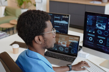 Fototapeta na wymiar Young black man in eyeglasses looking at graphs and charts on computer screens while sitting by workplace in office and analyzing data