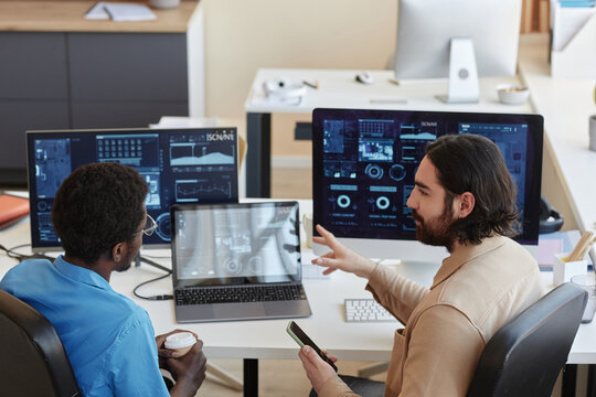 Young confident IT engineer explaining graphic data on computer screens to his colleague while looking at him during working meeting
