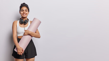 Horizontal shot of cheerful dark haired woman holds rolled karemat prepares for fitness training poses with headphones around neck dressed in cropped top and black shorts isolated on white background