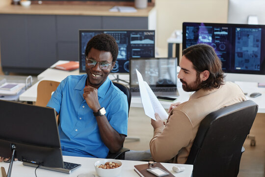 Happy young male analyst and his colleague looking through data on computer screen while one of them making presentation at meeting