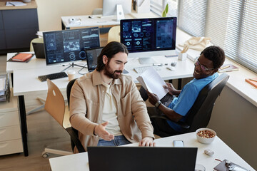 Young businessman showing data on computer screen to smiling African American colleague while explaining him results of analyzing it
