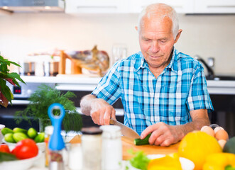 elderly man cuts vegetables for salad at the table in the kitchen