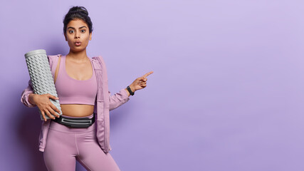 Photo of surprised Iraninan sportswoman carries massage roller has stunned expression points finger at empty space for your advertisement dressed in sportswear isolated over purple background.