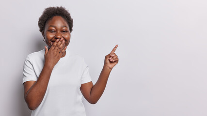 Dark skinned woman giggles positively covers mouth with hand points index finger at empty blank space shows something funny dressed in casual t shirt isolated over white background. Look at this