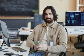 Young bearded IT engineer with smartphone texting and looking at camera while sitting by workplace...