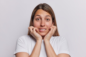 Nervous worried long haired European woman bites lips keeps hands on cheeks worries before important event dressed in casual t shirt isolated over white background hears unexpected bad news.