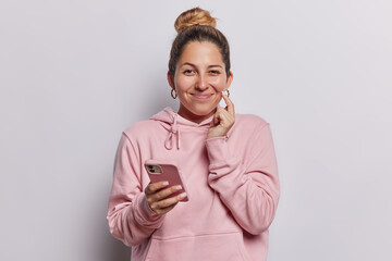 Cheerful pleased young woman holds mobile phone has cunning expression intention to do something wears casual pink sweatshirt isolated over white background. People and modern technologies concept