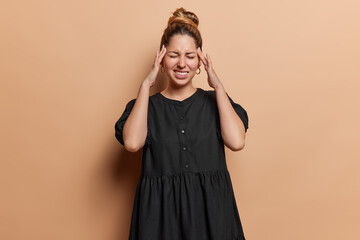 Indoor shot of pretty young woman suffers from migraine rubs her temples keeps hands on temples has eyes closed clenches teeth to reveal pain wears black dress isolated over brown background.