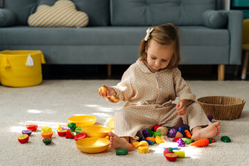 A little girl playing with wooden fruits and vegetables  on floor  in nursery. Educational games,...