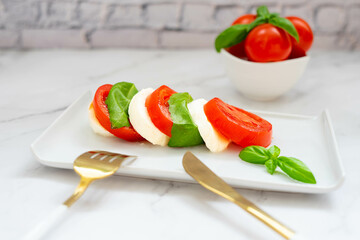 Caprese salad with mozzarella, tomatoes and fresh basil, on a white ceramic plate,  with white golden fork and knife, on a marble background. Italian cuisine