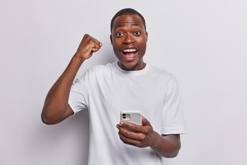 Confident happy dark skinned man holding mobile phone and celebrating success clenches fist exclaims like winner glad to win lottery dressed in casual t shirt isolated over white background.