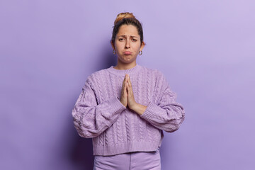 Uneasy dissatisfied woman upset and seeking mercy apologizes and pleads for help pressing her palms together in praying gesture begs for favor wears casual knitted jumper isolated on purple background