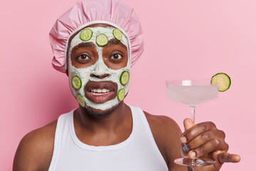 Displeased African man taking spa procedure in wellness center applies clay mask with cucumber slices holds cocktail wears bath hat and white t shirt isolated over pink background. Mens cosmetology