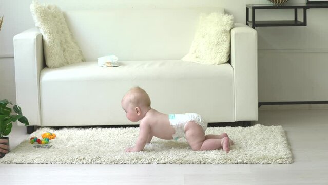 A healthy caucasoid baby lies on a white carpet on the floor in a bright room to learn to crawl indoors. An adorable baby keeps his belly and feet on the floor to crawl on his belly fun and happily at