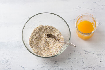 A glass bowl with ground oatmeal and a glass of freshly squeezed orange juice on a light gray background, top view. Cooking a healthy homemade breakfast