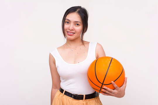 A smiling young Asian woman posing while holding a basketball in hand. Isolated on a white background.