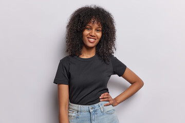 Cheerful self confident African woman keeps hand on waist smiles gladfully wears casual black t shirt and jeans poses against white studio background enjoys spare time. People and emotions concept